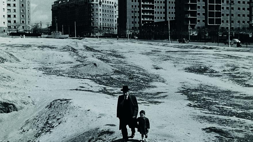 Fotografía en blanco y negro de un hombre y su pequeño nieto paseando por un descampado y, al fondo, una serie de edificios de la periferia de Madrid