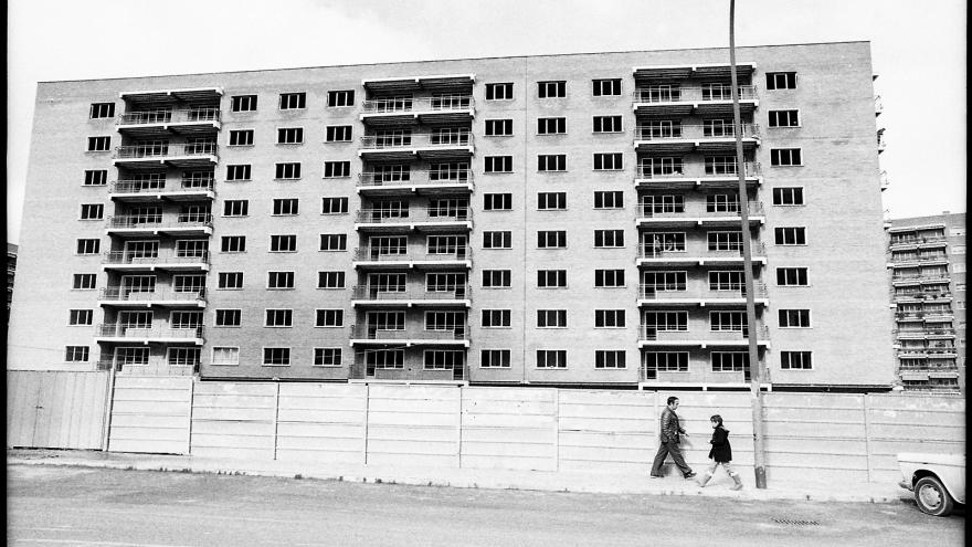 Fotografía de Javier Campano, edificio rodeado por una valla de obras, un hombre y un niño se cruzan por la acera en el barrio de Hortaleza