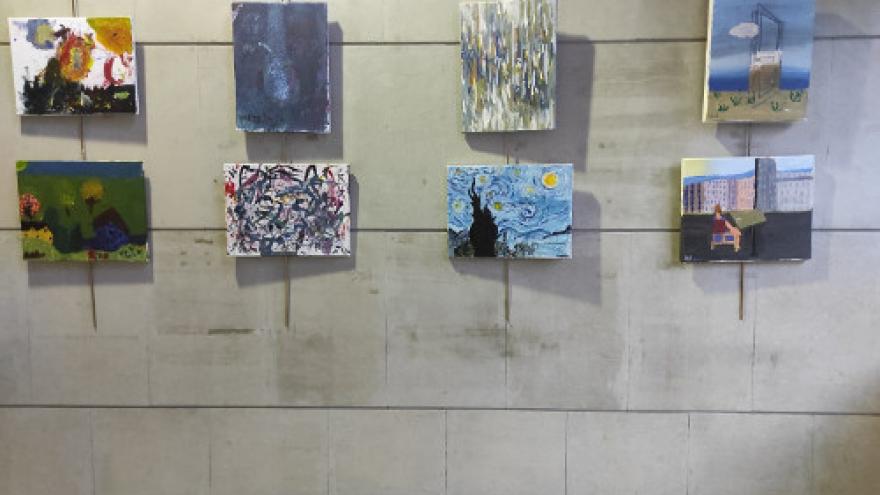 Fotografía de una pared gris , cuelgan lienzos realizados por los alumnos y alumnas del taller 