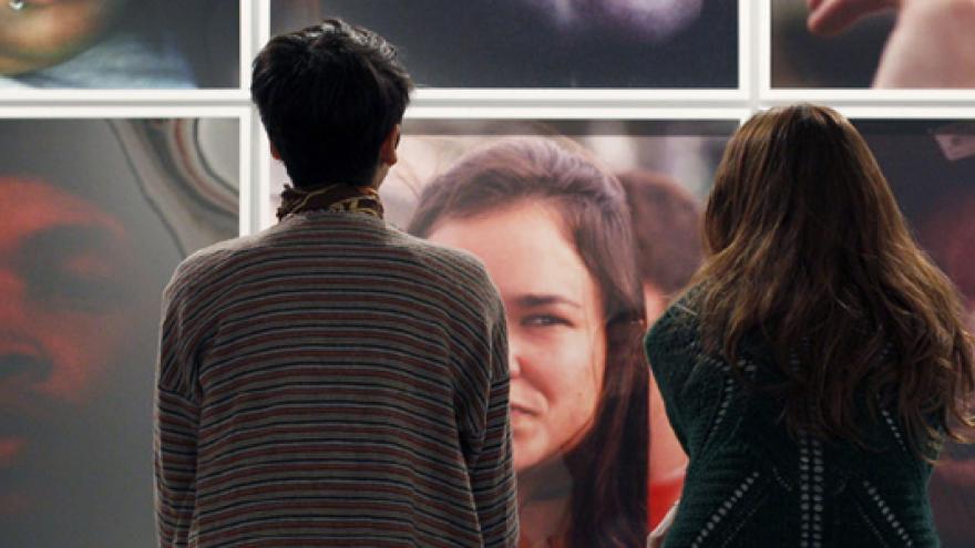 Dos mujeres sentadas de espaldas al espectador y mirando una pared llena de fotografías con retratos en color de personas anónimas