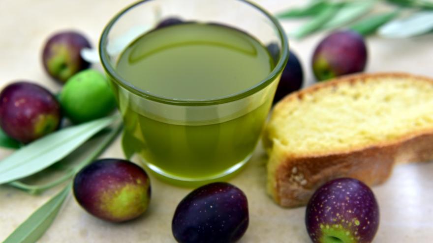 Vaso de aceite de oliva junto a una rebanada de pan y aceitunas alrededor