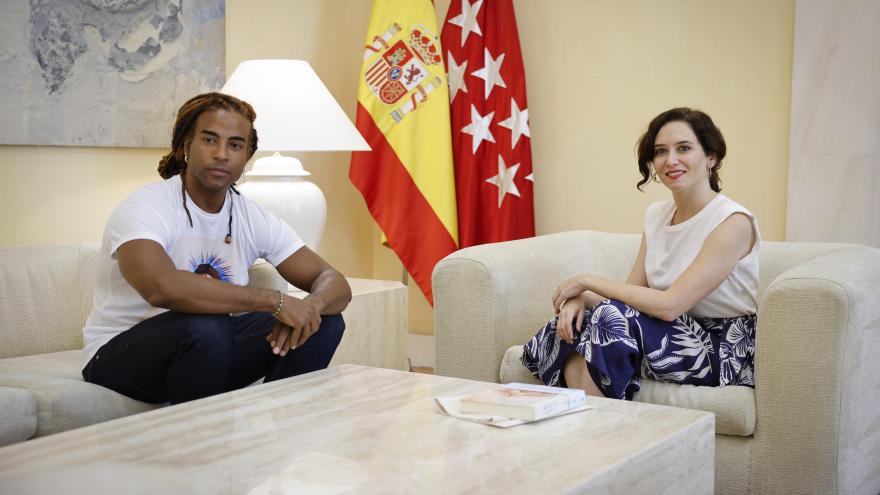 El artista Yotuel sentado a la izquierda de la presidenta en unos sofás con una mesa en medio
