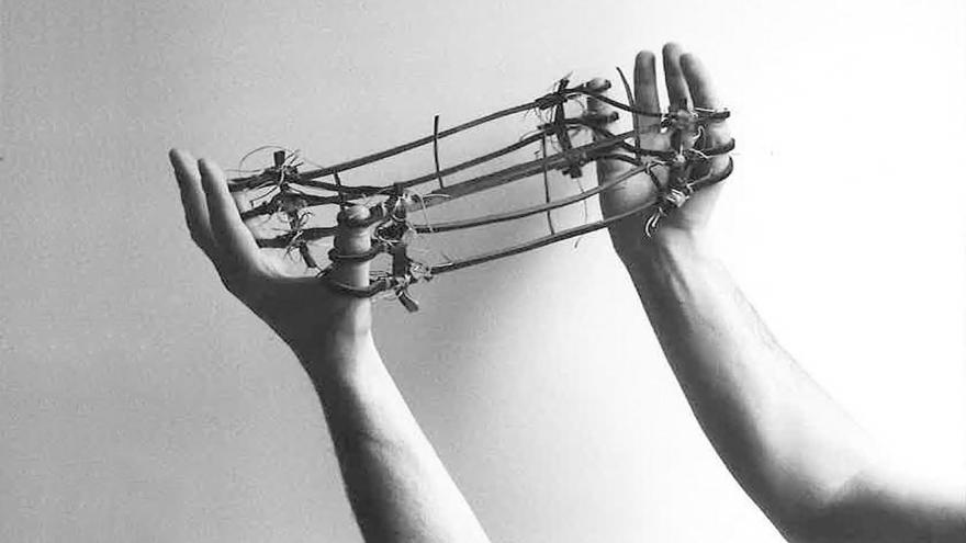 Fotografía en blanco y negro que muestra dos brazos y varias cuerdas con espinas entrelazadas en los dedos de ambas manos