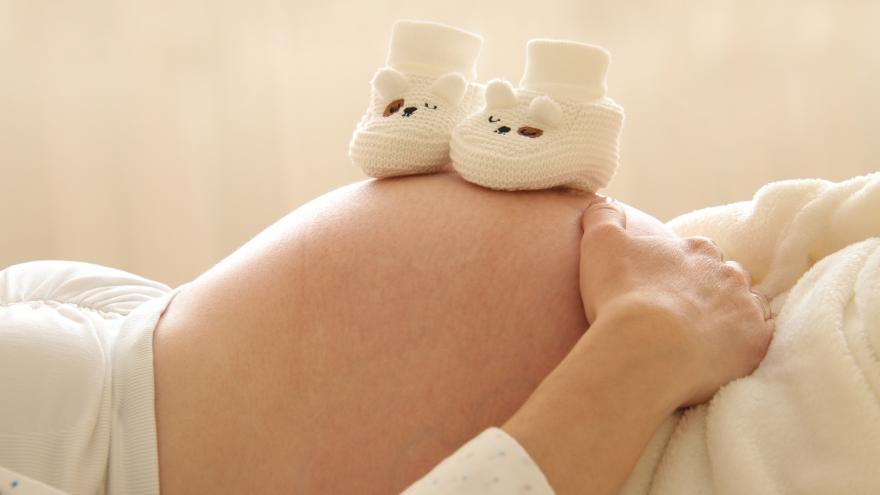 Sobre el vientre de una mujer embarazada que está tumbada bocarriba hay dos patucos de bebé