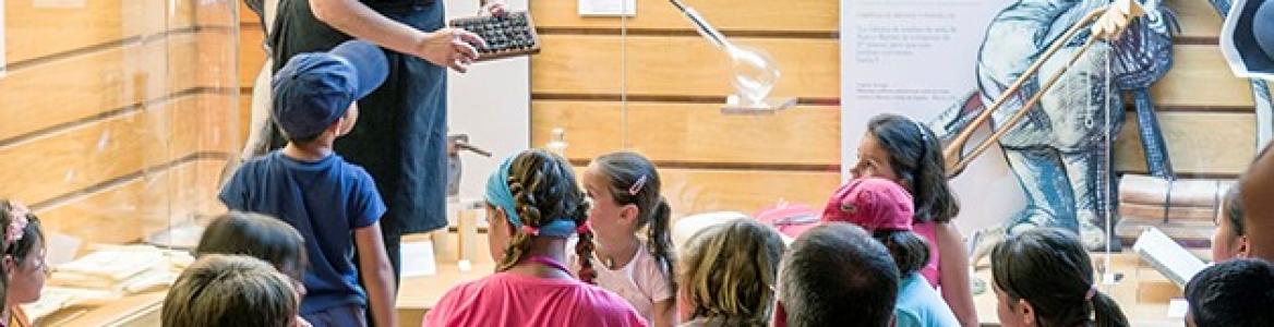 Niños y niñas en el centro de interpretación mirando a la educadora de la actividad que les explica cómo se hace el vídrio
