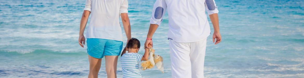 Familia en la playa cogidos de la mano. El bebé, en medio, sujeta un peluche