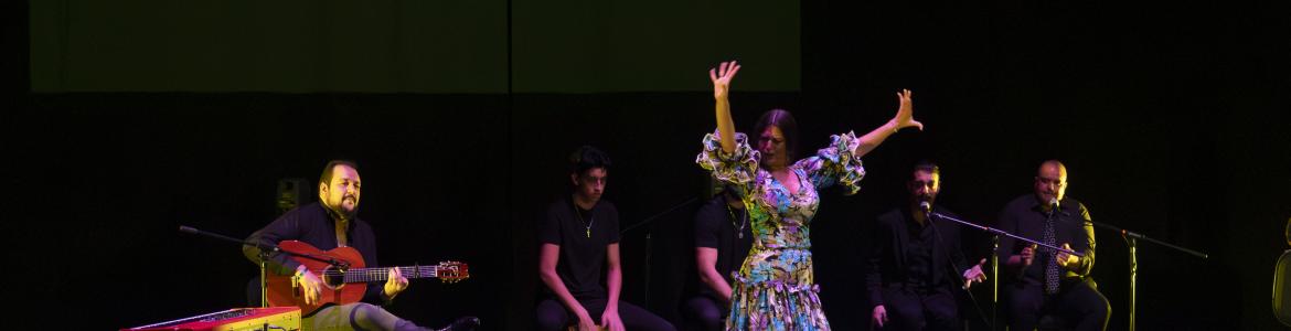 mujer con vestido con volantes baila con los brazos en alto, en el fondo un grupo de músicos con guitarra y cajón flamencos