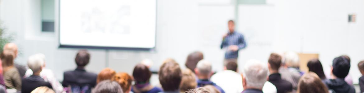 Un grupo de personas sentadas en un aula, escuchando a un conferenciante que está al fondo, delante de una pared y de una pizarra