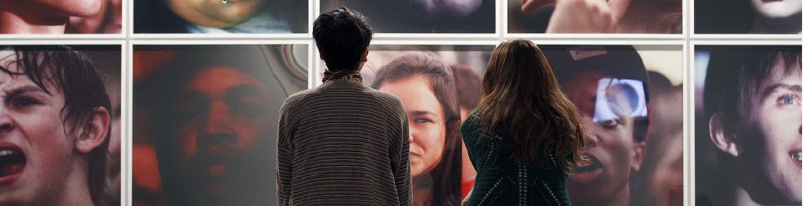 Dos mujeres sentadas de espaldas al espectador y mirando una pared llena de fotografías con retratos en color de personas anónimas