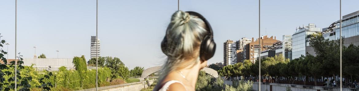 Mujer escuchando música con cascos, girada al espectador, en un parque rodeado de edificios