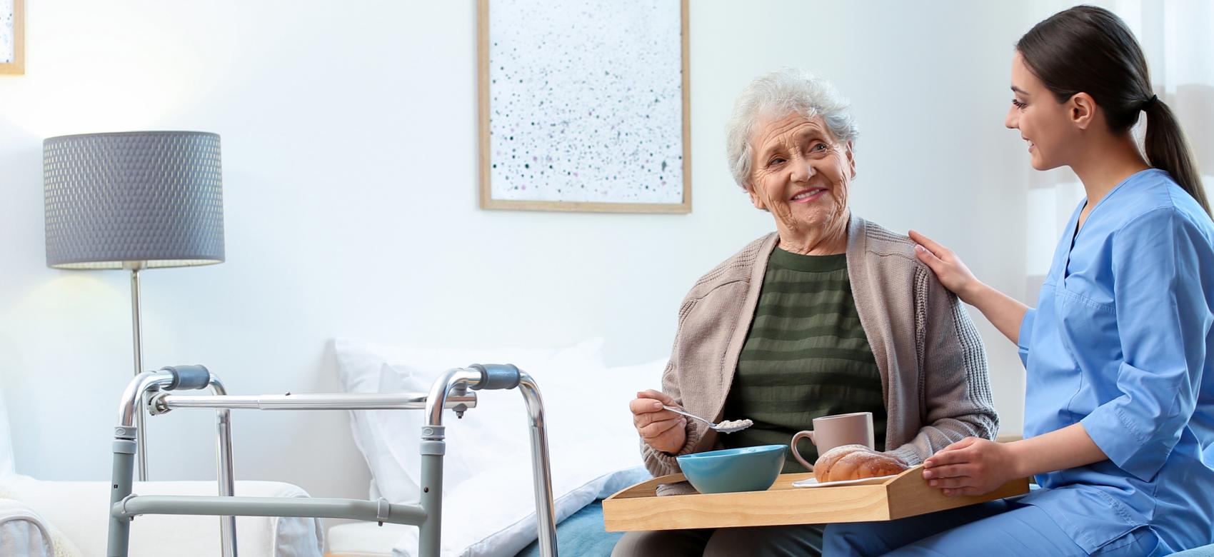 Coordinación sociosanitaria Una persona mayor en una habitación de un centro residencial comiendo acompañada de una profesional sanitaria