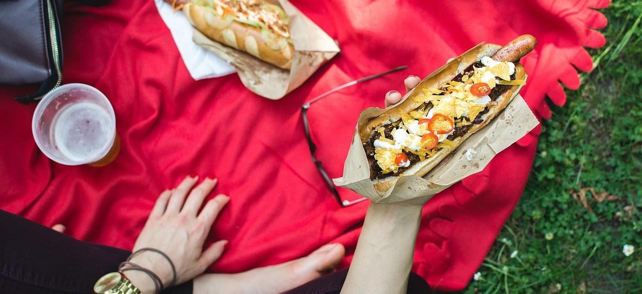 Picnic en la hierba sobre mantel rojo y mano agarrando un perrito caliente