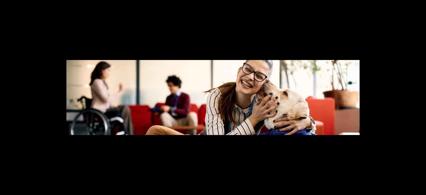 Persona con discapacidad abraza a un perro de asistencia en una oficina con otras personas conversando al fondo