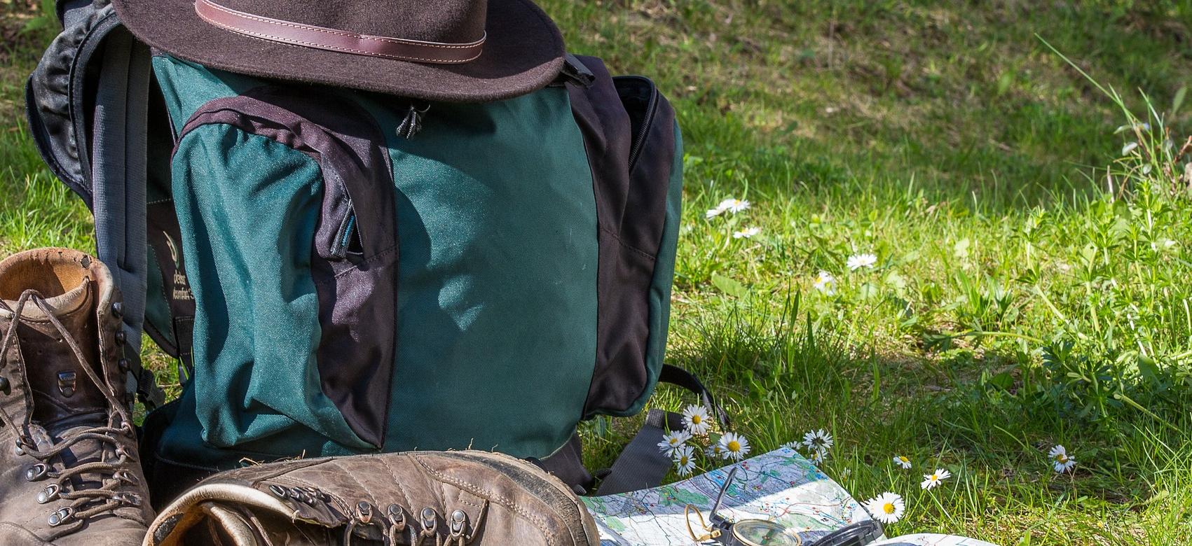 Unas botas de senderismo marrores, un plano, una brujula, una mochila de color azul y negra y encima de esta un sombrero de ala de color marrón oscur