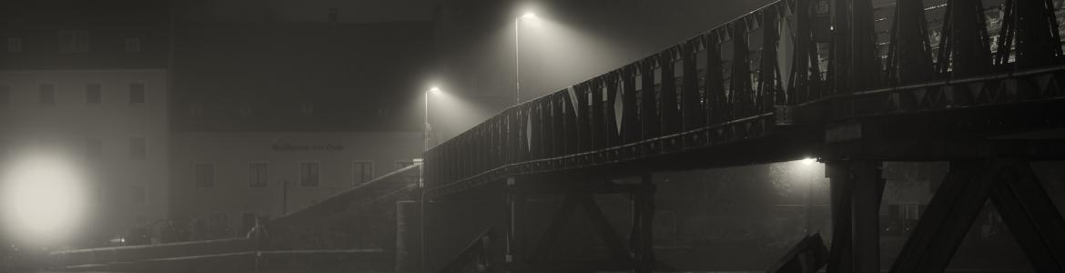 Imagen de un puente y casas al fondo, de noche, con bruma