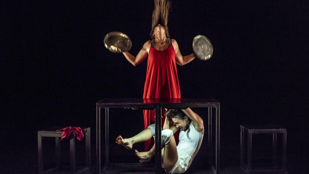 Dos mujeres hacen una performance. Una de ellas está encerrada en una caja de cristal y la otra, vestida de rojo, toca unos platillos de metal.