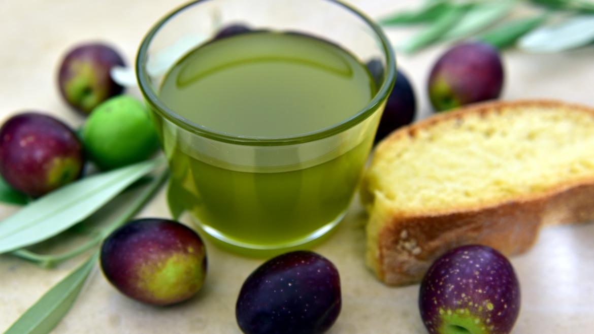 Vaso de aceite de oliva junto a una rebanada de pan y aceitunas alrededor