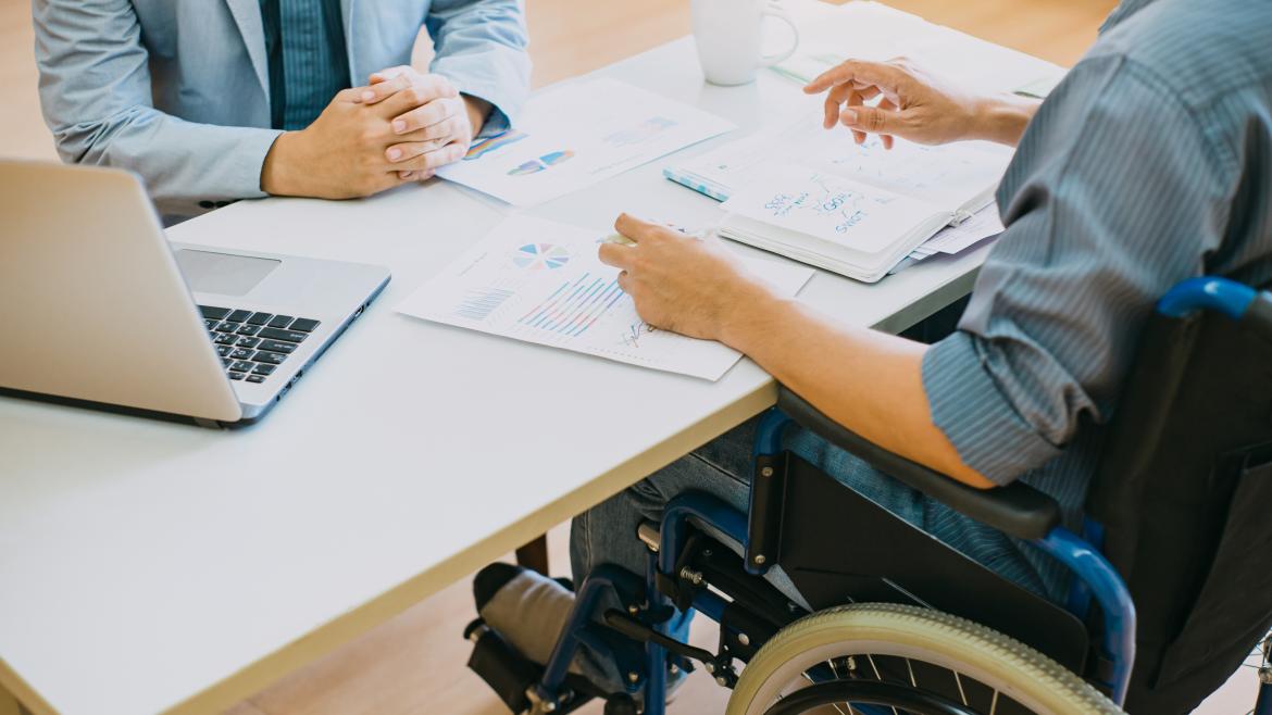 Persona en silla de ruedas sentado en una mesa con otra persona y muchos papeles con gráficos en la mesa