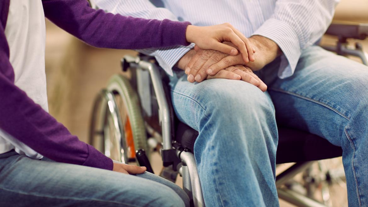 Una mujer pone sus manos sobre las de un hombre que está en una silla de ruedas