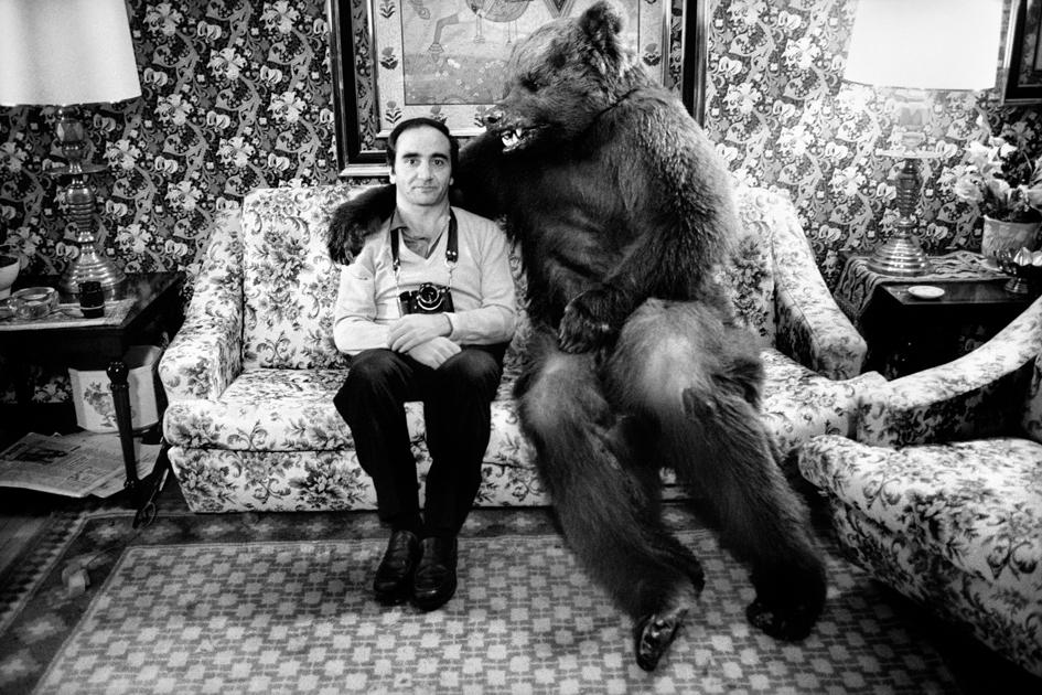 Hombre sentado en un sofá junto a un oso pardo que le abraza con su garra sobre el hombro