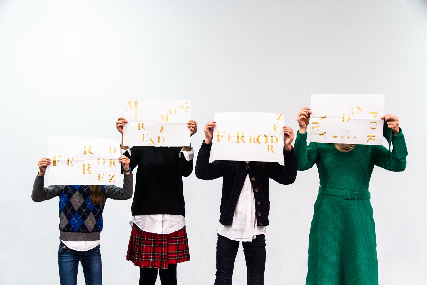 Cuatro personas, mujeres, niños y niñas, fotografiados de frente mientras se tapan su rostro con un folio lleno de letras doradas