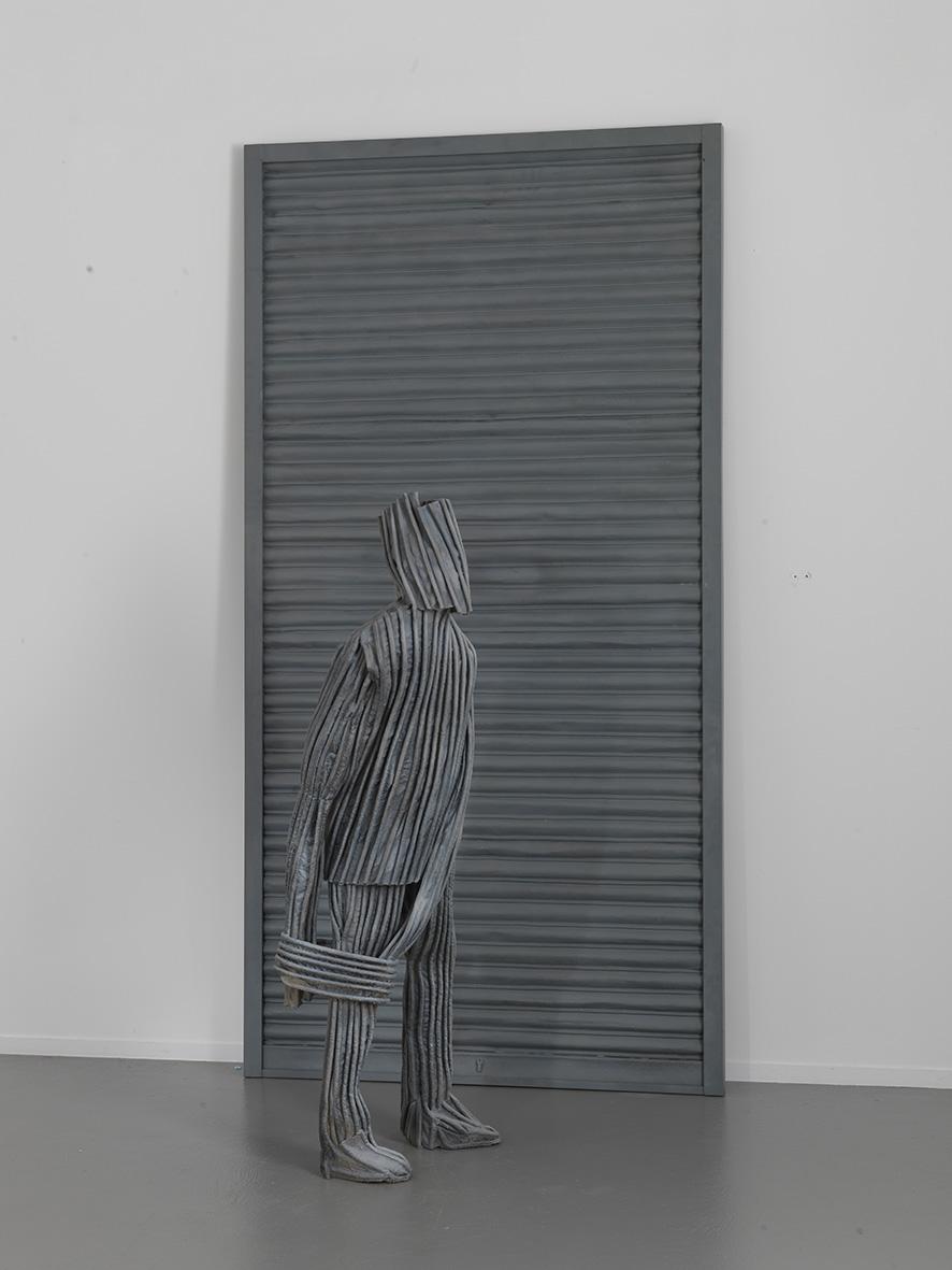 Sculpture of a man with a covered head and on a grooved wall, like his clothes