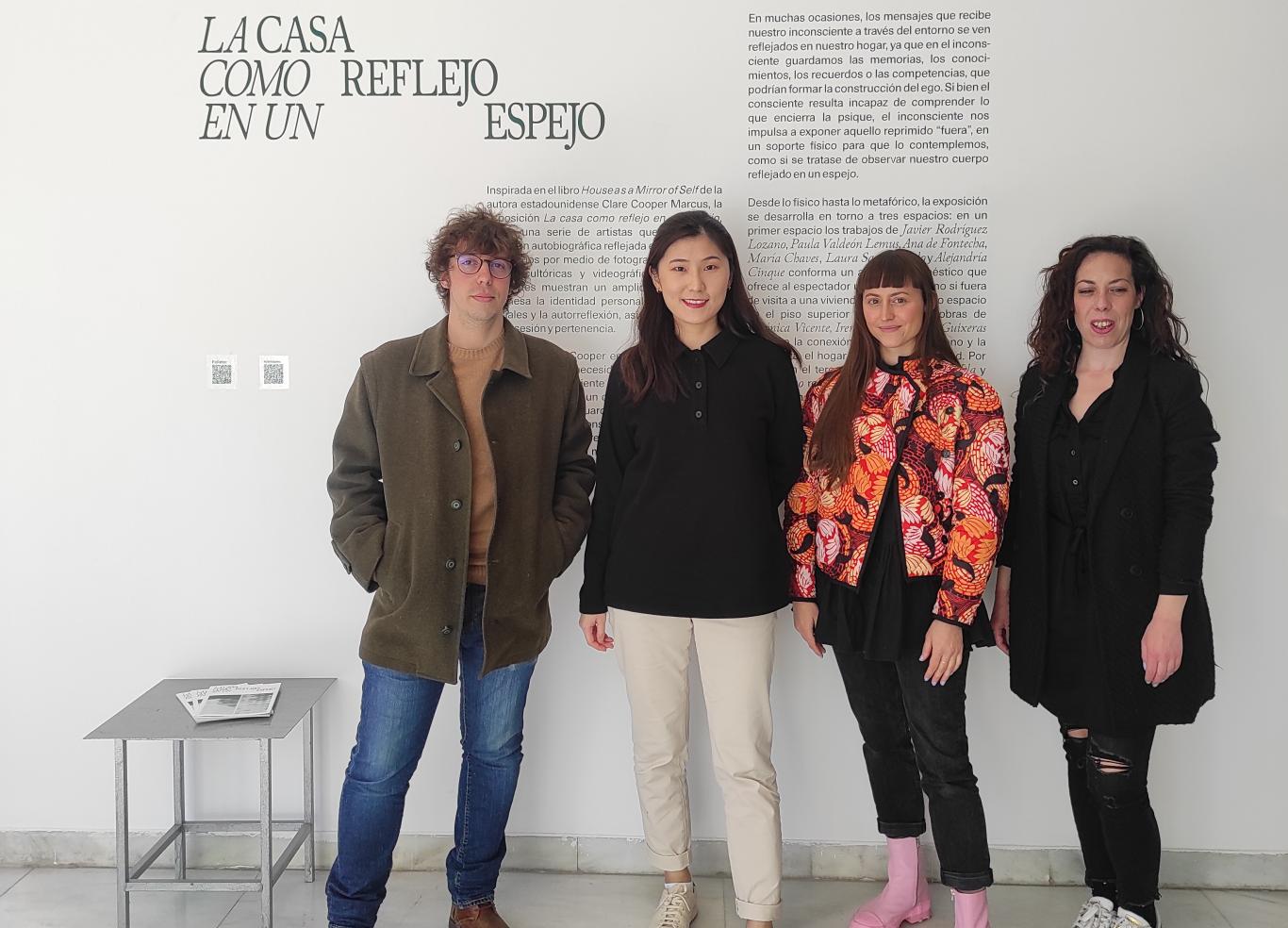 Cuatro personas jóvenes posando ante la cámara frente al titulo de una exposición que se llama la casa como reflejo en un espejo
