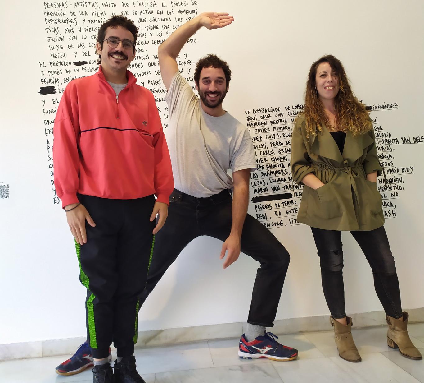 Dos hombres jóvenes y una mujer posando frente al cartel de la exposición Licra