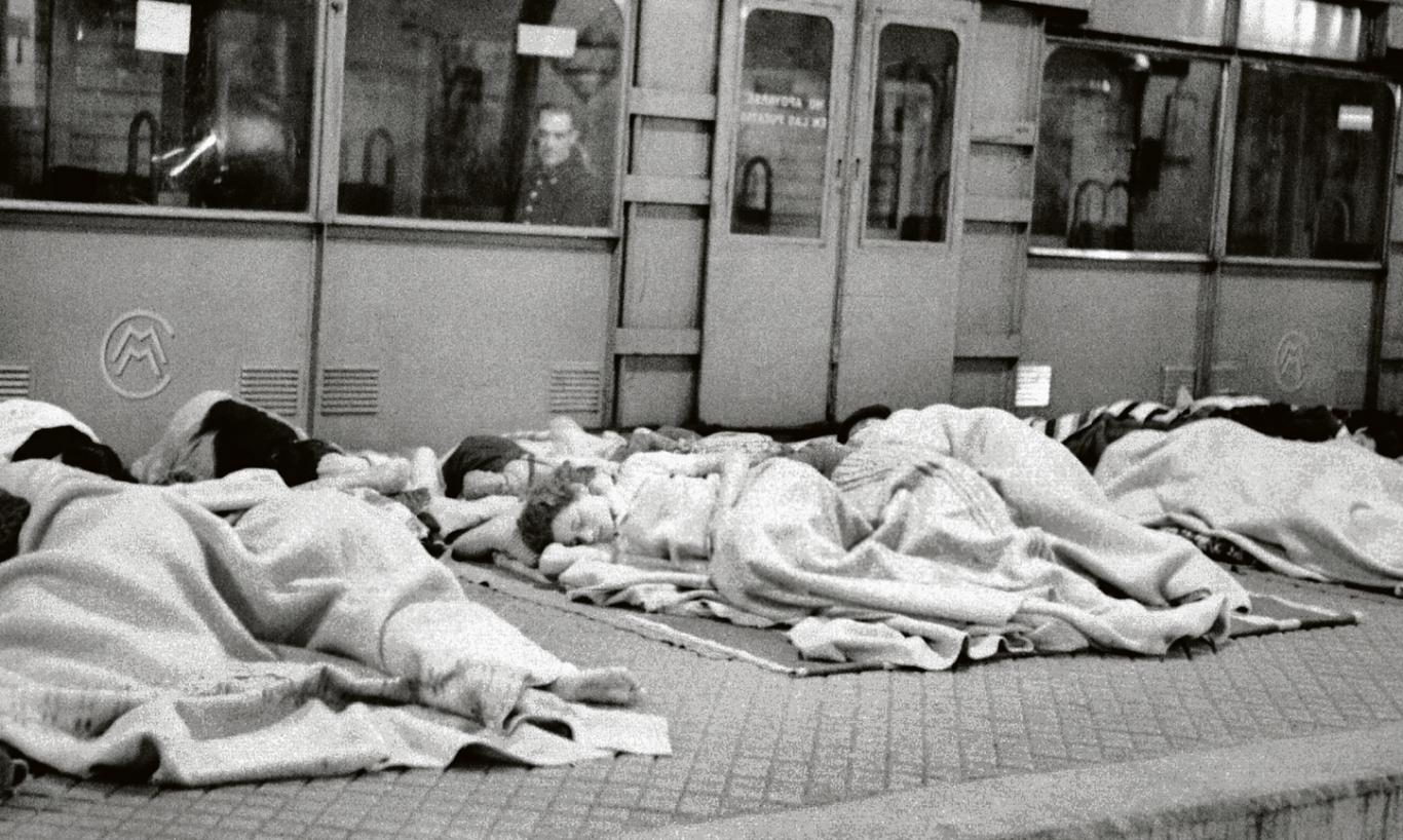 Varias personas duermen en el andén del metro mientras un hombre uniformado les observa desde el interior de un vagón