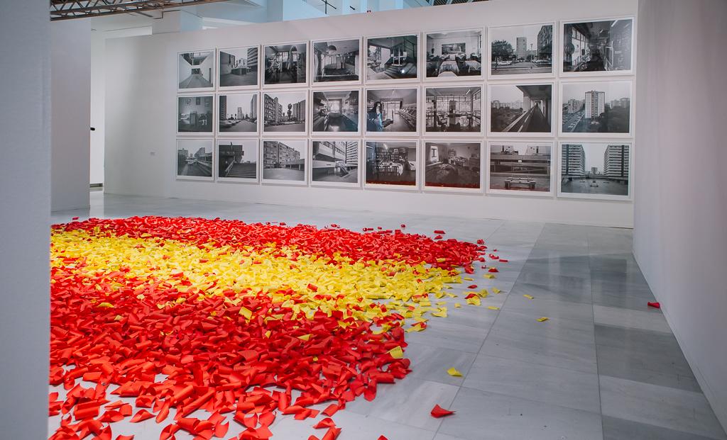 Instalación artística con papeles de colores tirando en el suelo que conforman, por agrupación, la bandera de España. En el fondo una composición fotográfica