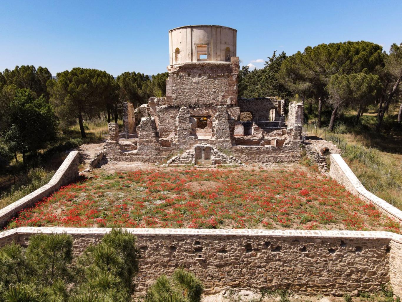 edificio con torreón semicircular y perímetro de piedra, rodeado por un pinar y un campo de amapolas