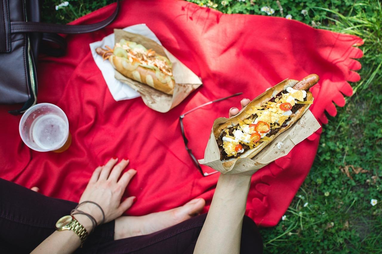 Picnic en la hierba sobre mantel rojo y mano agarrando un perrito caliente