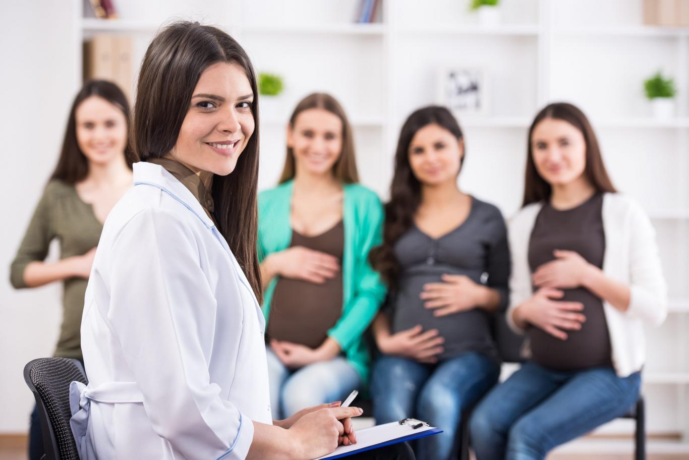 En primer plano una joven doctora sonriente y en segundo término desenfocadas cuatro mujeres jóvenes embarazadas tocándose el viente