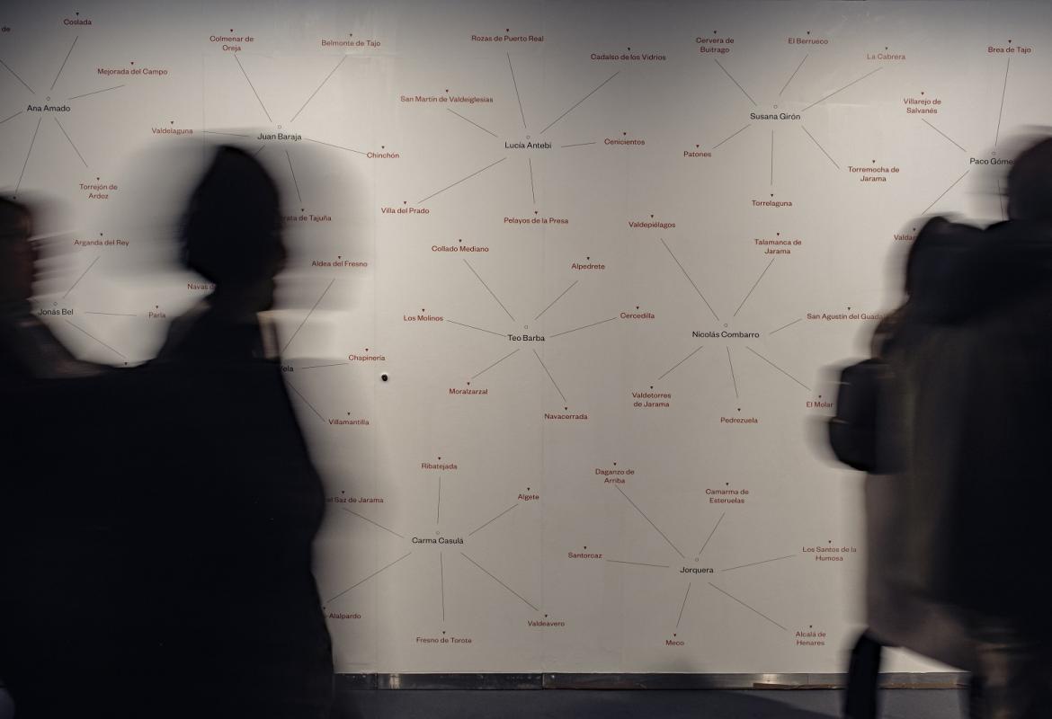 Muro de la exposición en el que aparecen los nombres de los fotógrafos y de los municipios, se aprecian sombras de personas